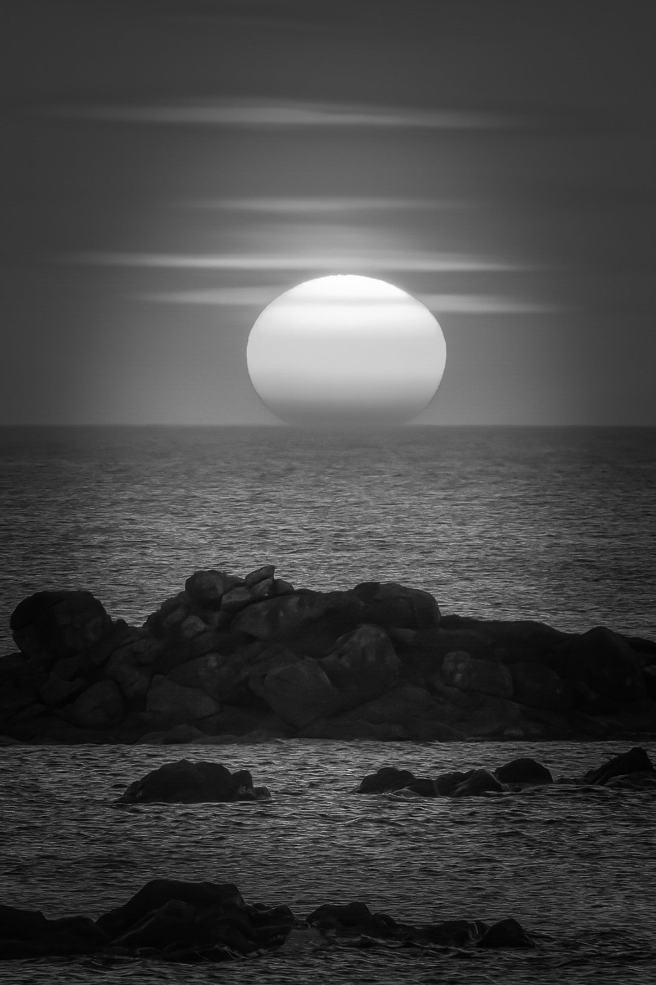 Soleil couchant au centre de l’image, mer bleue et rochers, reflet rouge et orange du ciel sur l’eau, noir et blanc