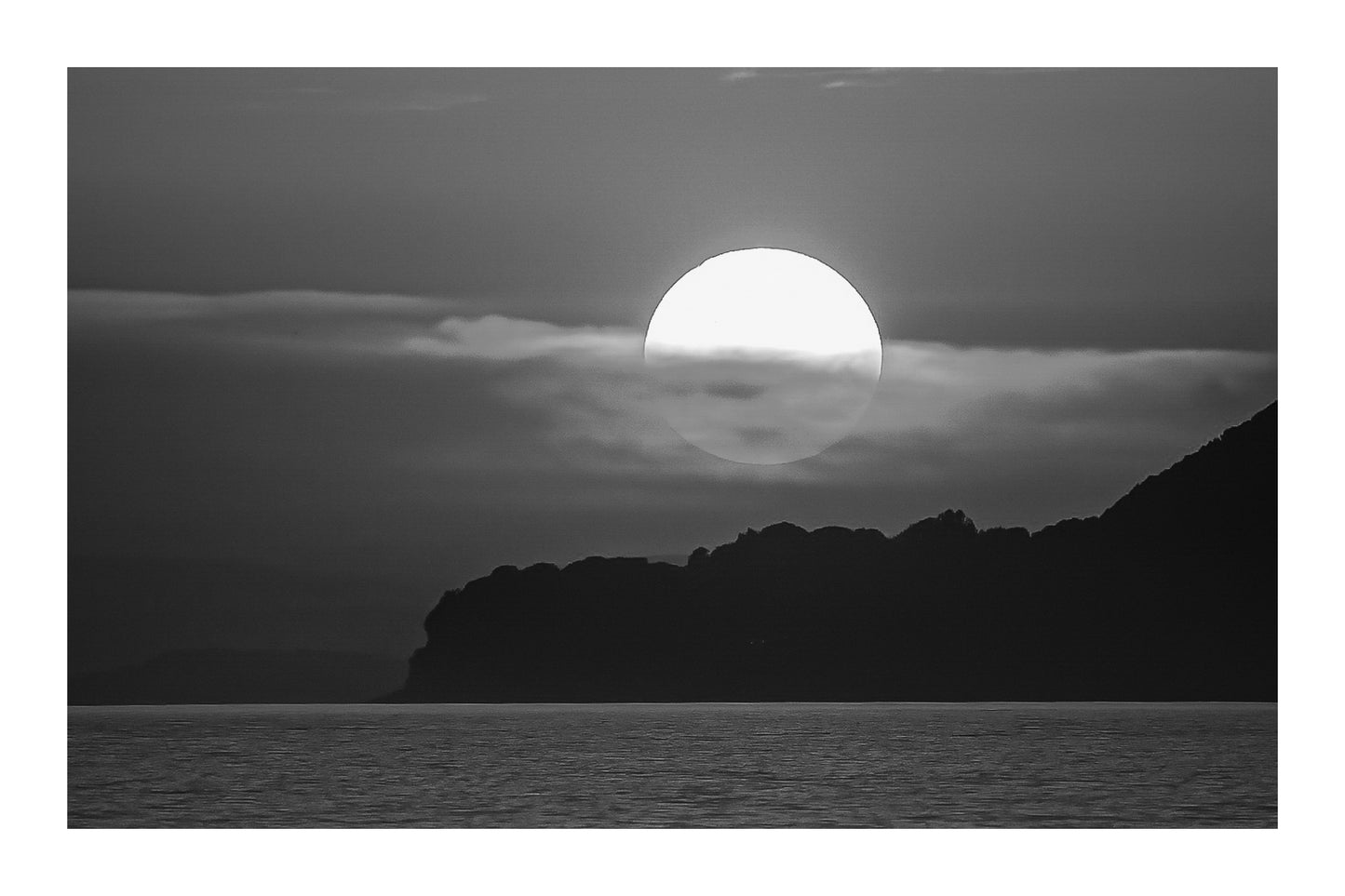 Gros plan sur le soleil rougeoyant partiellement voilé par un nuage au-dessus de la silhouette sombre de Giens, noir et blanc avec bordure