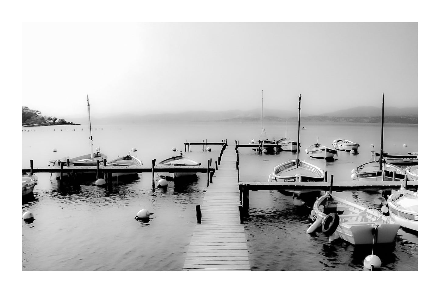 Ponton en bois menant à une rangée de barques, mer lisse et brume douce au port du Brusc, noir et blanc avec bordure
