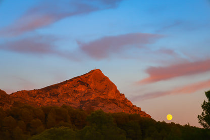 Lune jaune se levant près de la montagne Sainte-Victoire, ciel bleu aux nuages roses, pins en premier plan au crépuscule