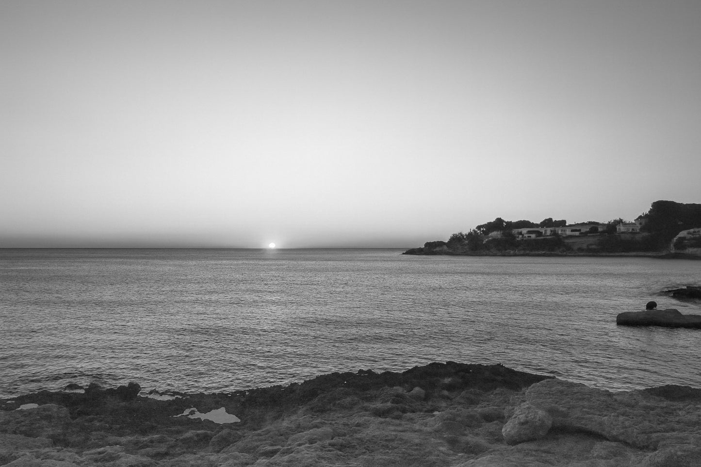 soleil à l’horizon éclairant une anse rocheuse et le village de Carry-le-Rouet, noir et blanc