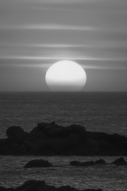 Soleil couchant parfaitement rond au-dessus de la mer, ciel orange strié de nuages et rochers au premier plan, noir et blanc