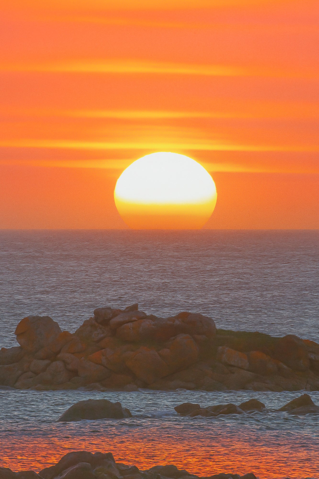 Soleil couchant parfaitement rond au-dessus de la mer, ciel orange strié de nuages et rochers au premier plan