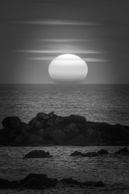 Soleil couchant au centre de l’image, mer bleue et rochers, reflet rouge et orange du ciel sur l’eau, noir et blanc