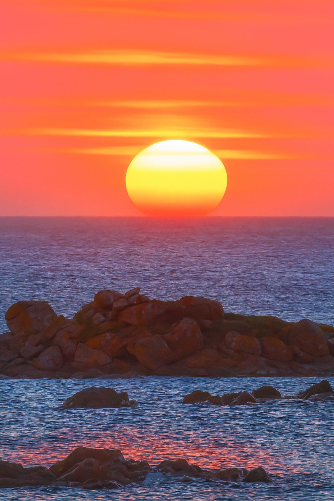 Soleil couchant au centre de l’image, mer bleue et rochers, reflet rouge et orange du ciel sur l’eau