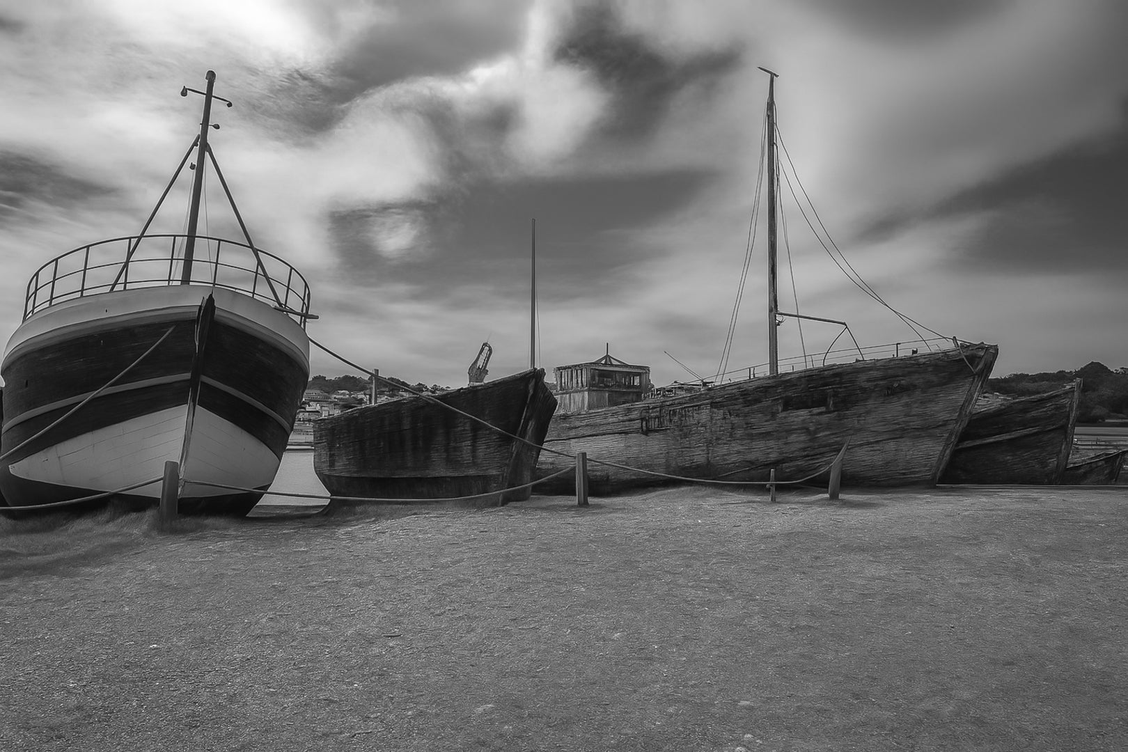 Trois bateaux en bois alignés sur la grève à Camaret, ciel nuageux et port en arrière-plan, noir et blanc