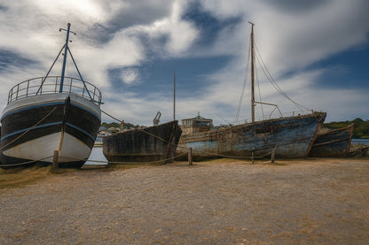 Trois bateaux en bois alignés sur la grève à Camaret, ciel nuageux et port en arrière-plan