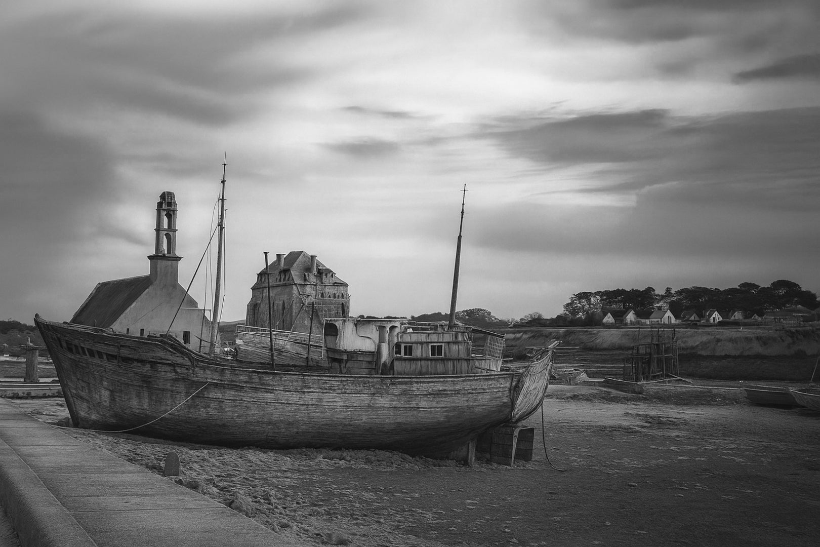 Épave bleue au premier plan devant chapelle et maisons, ciel doré au-dessus du port de Camaret, noir et blanc