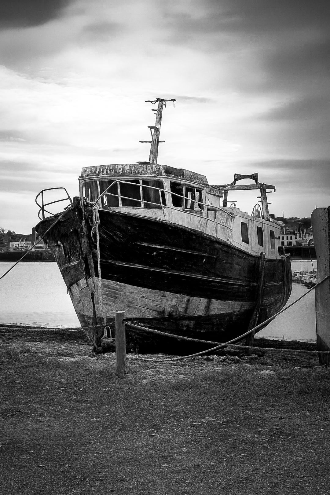 Épave au nez bleu et blanc amarrée par des cordes sur l’estran, ciel couvert au port de Camaret, noir et blanc