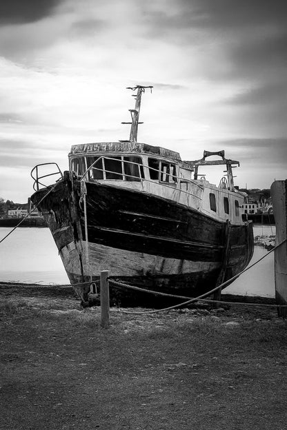 Épave au nez bleu et blanc amarrée par des cordes sur l’estran, ciel couvert au port de Camaret, noir et blanc