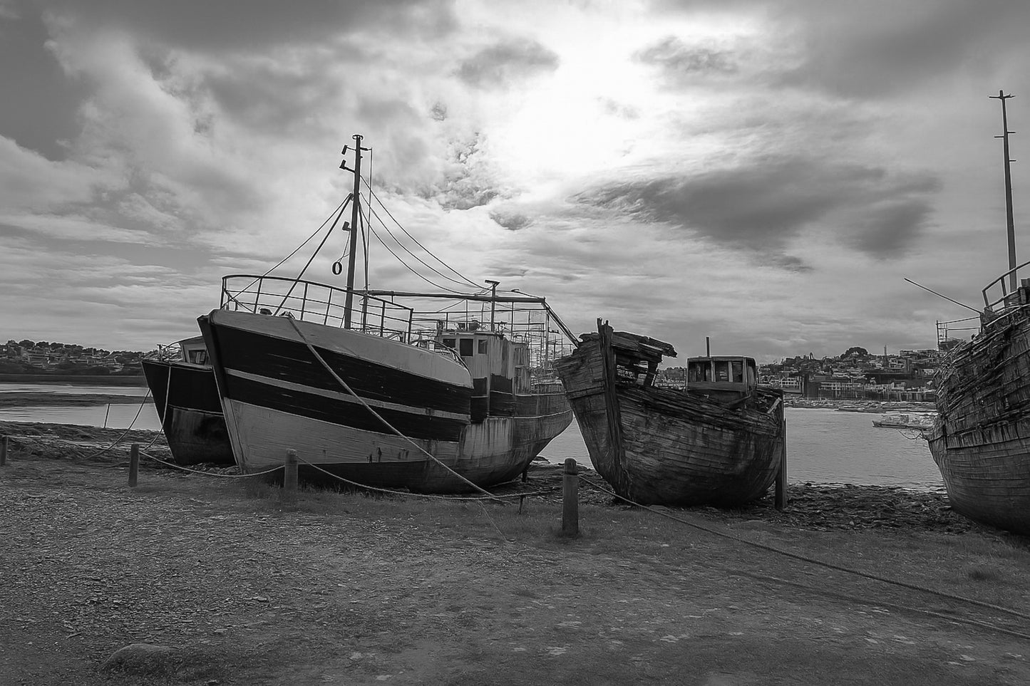 Bateaux récents à gauche et coque déchiquetée à droite sur la grève de Camaret, ciel lumineux, noir et blanc