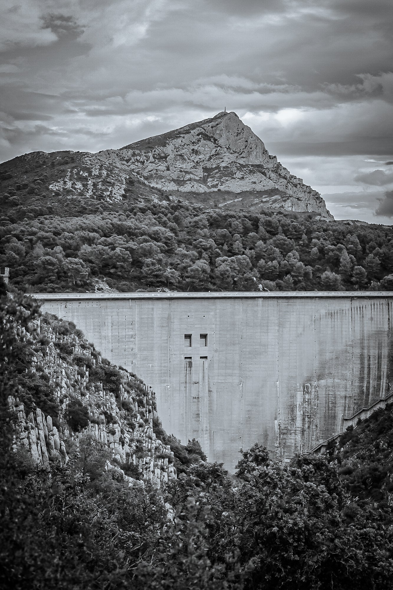 Barrage massif au premier plan avec la Sainte-Victoire en arrière-plan, forêt méditerranéenne et ciel nuageux, noir et blanc