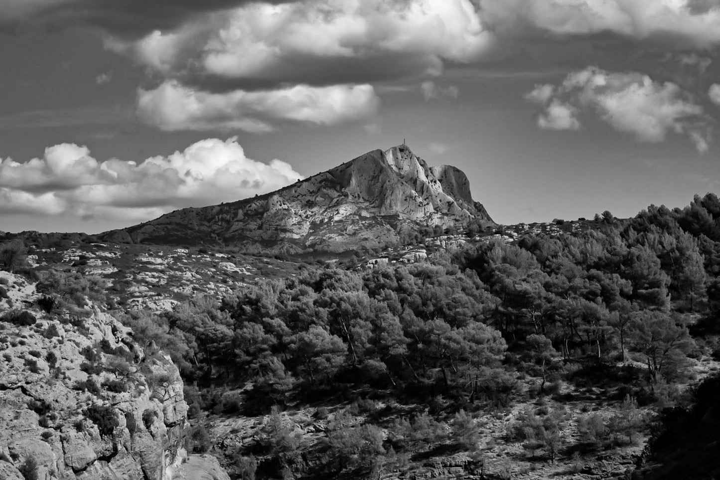 Montagne Sainte-Victoire se dressant au-dessus d’une forêt de pins, surmontée de gros nuages blancs, noir et blanc