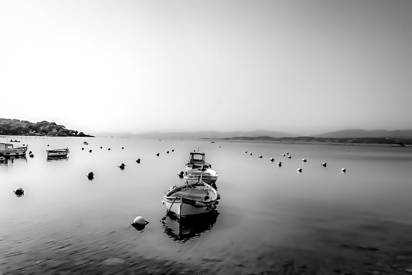 Trois barques alignées dans l’axe, bouées et eau immobile, brume et collines lointaines au Brusc, noir et blanc