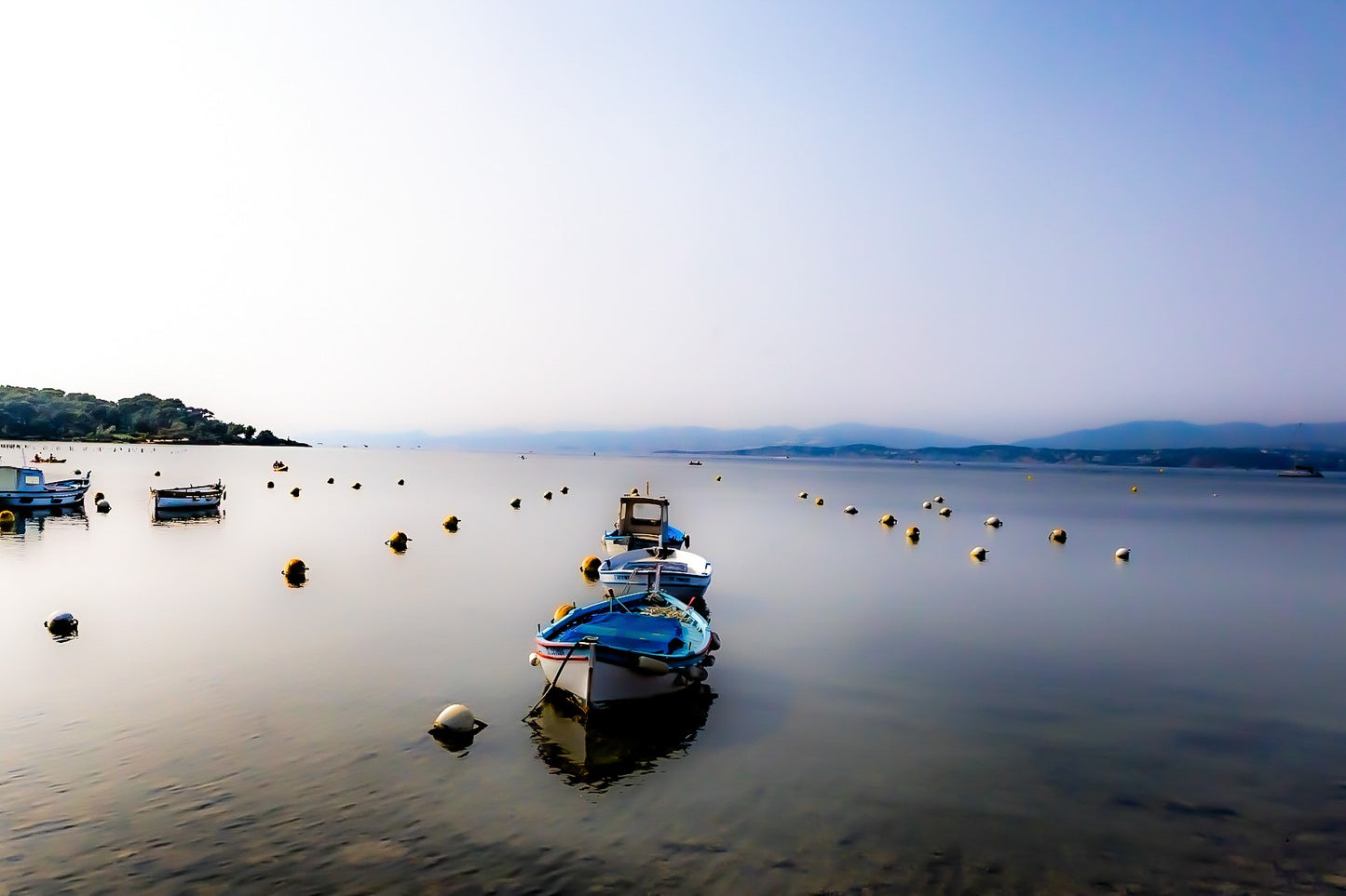 Trois barques alignées dans l’axe, bouées et eau immobile, brume et collines lointaines au Brusc, couleur
