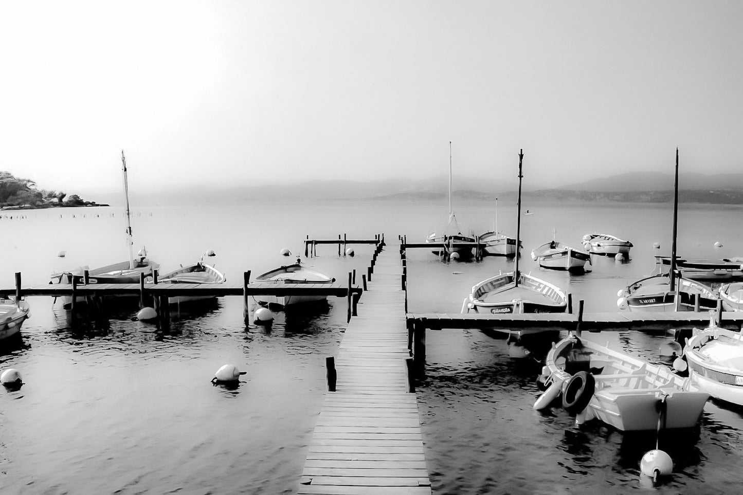 Ponton en bois menant à une rangée de barques, mer lisse et brume douce au port du Brusc, noir et blanc