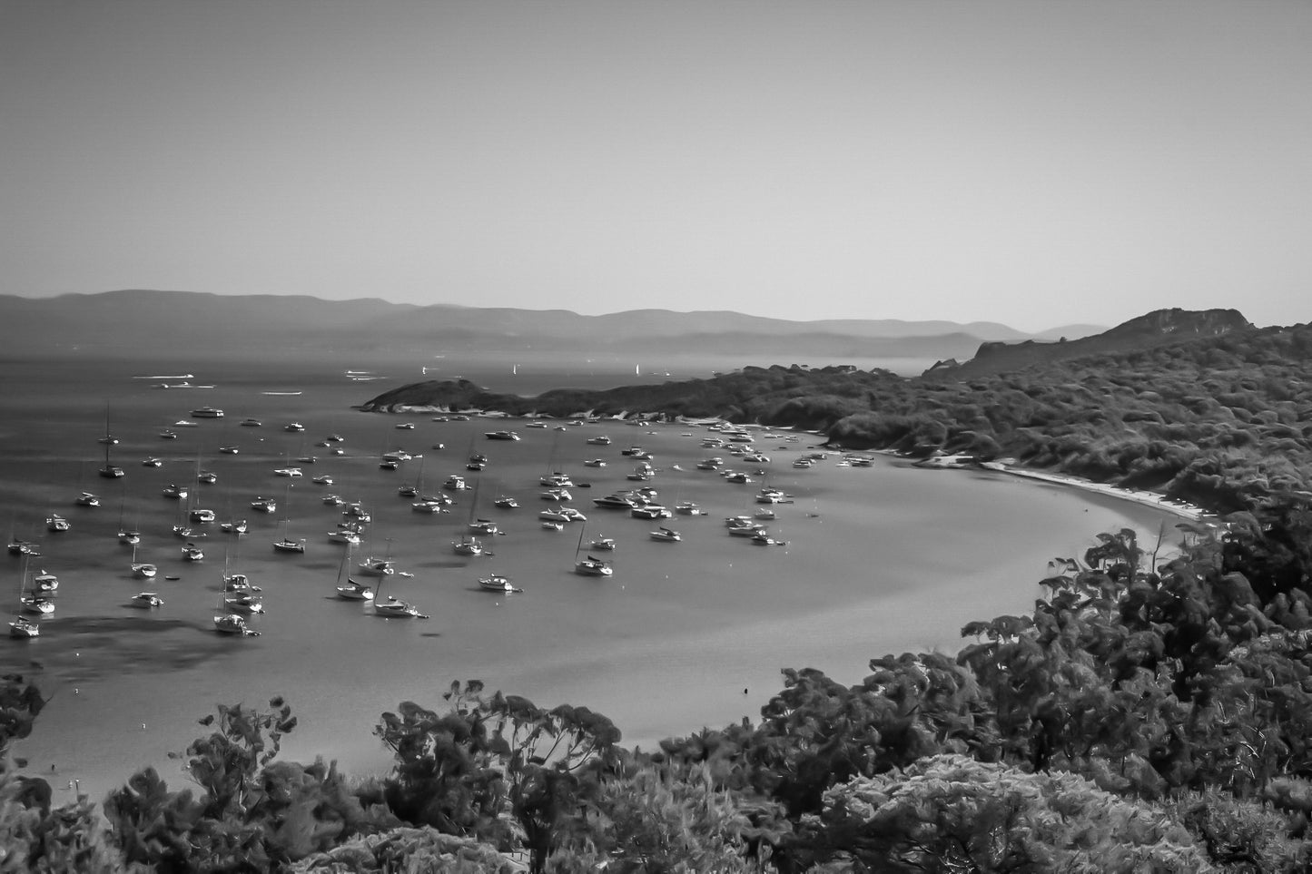 Vue plongeante sur la grande baie de Porquerolles avec de nombreux bateaux ancrés dans une eau turquoise sous un ciel d’été, noir et blanc