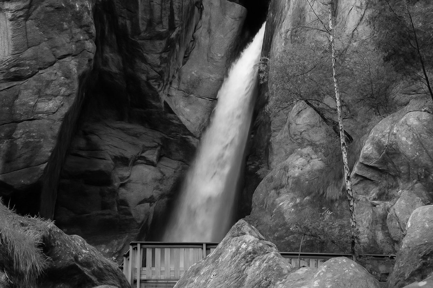 Grande chute d’eau de la cascade du Ray au fond d’une faille rocheuse, passerelle de bois au premier plan, noir et blanc