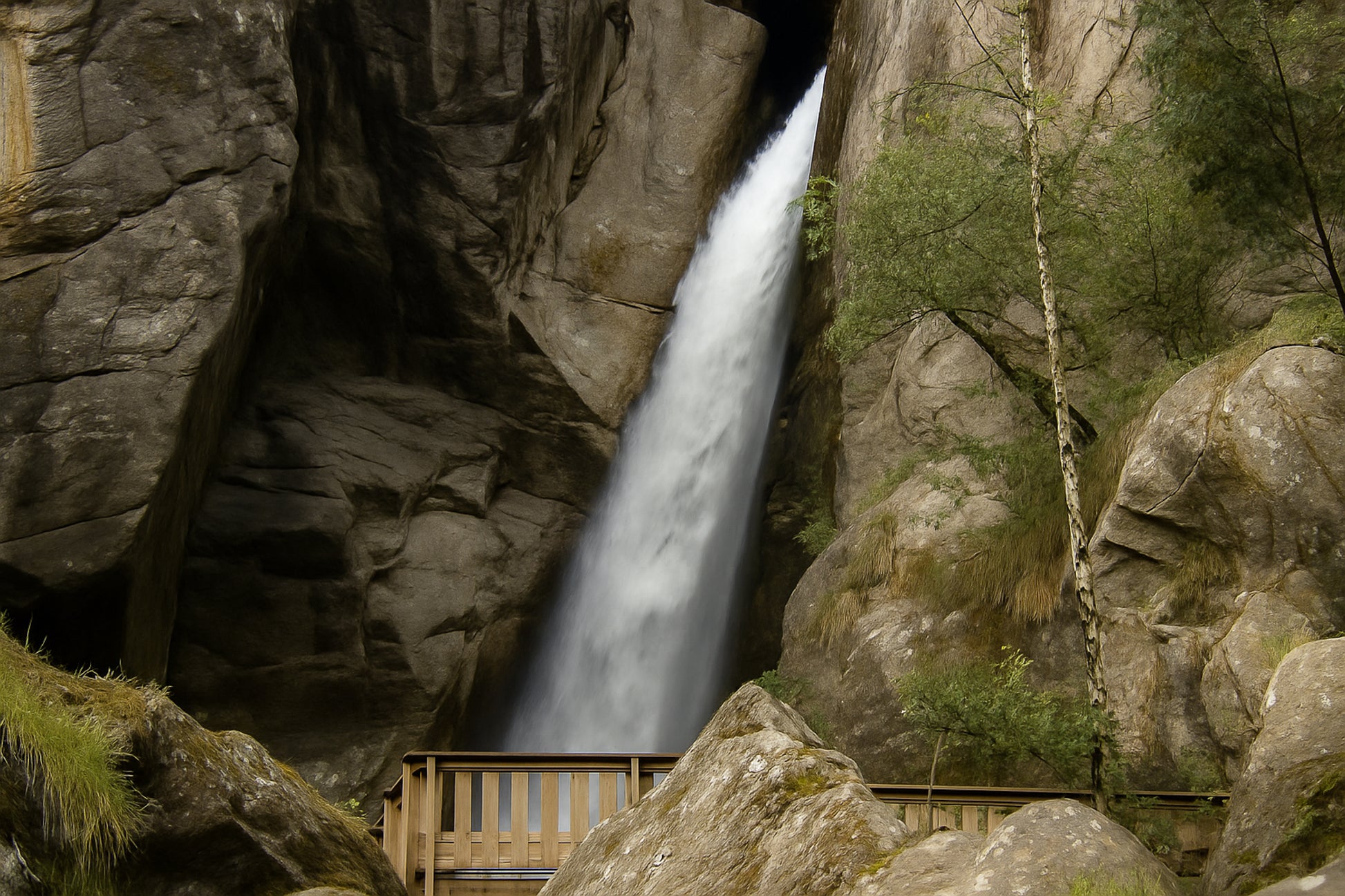 Grande chute d’eau de la cascade du Ray au fond d’une faille rocheuse, passerelle de bois au premier plan, couleur