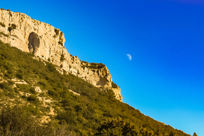 Falaise calcaire de la Sainte-Baume baignée de lumière chaude avec la lune accrochée dans un ciel bleu profond.