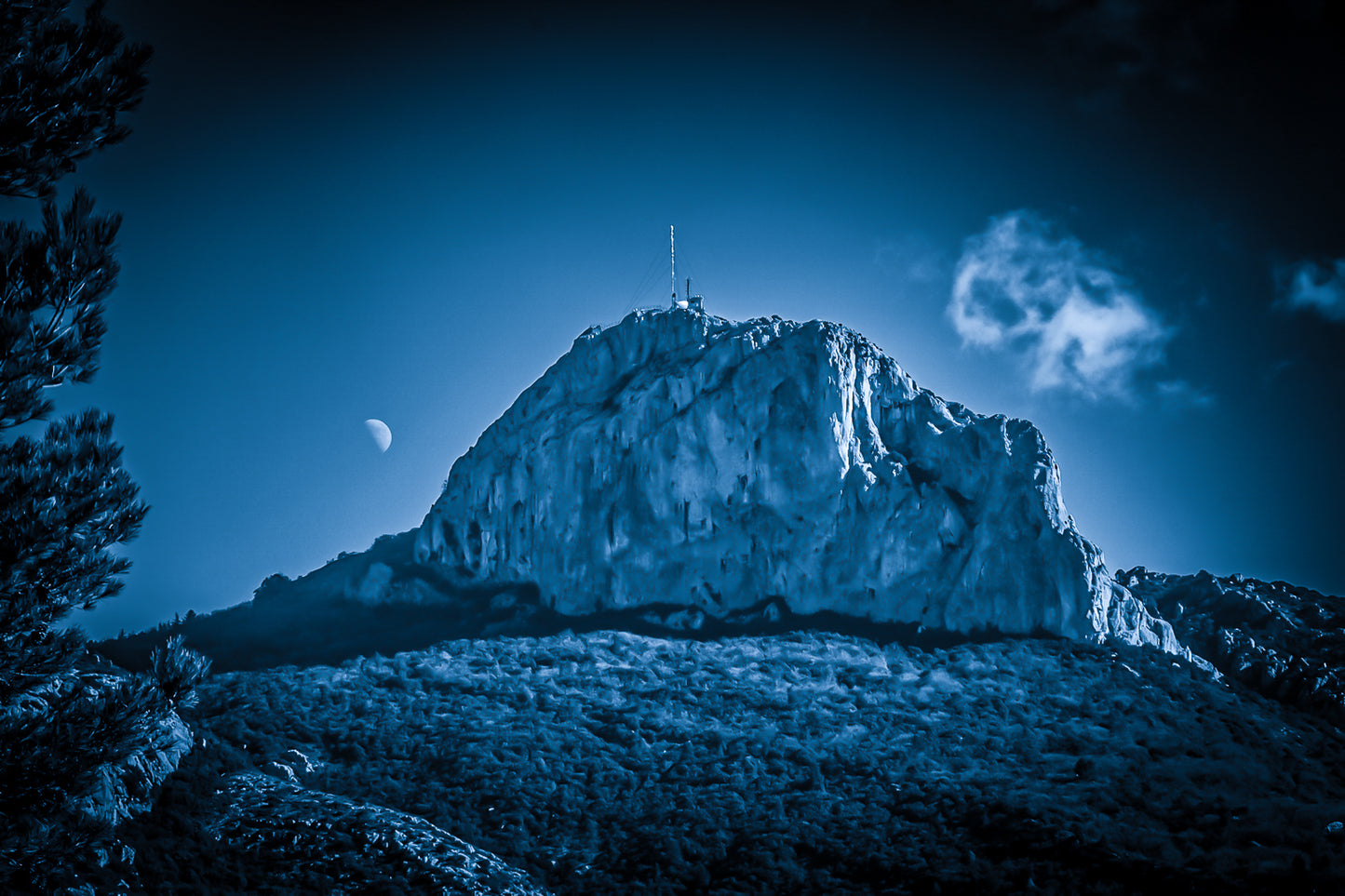 Massif de la Sainte-Baume baigné d’une lumière bleutée avec la lune au-dessus, nuages légers et forêt sombre.