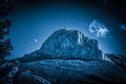 Massif de la Sainte-Baume baigné d’une lumière bleutée avec la lune au-dessus, nuages légers et forêt sombre.