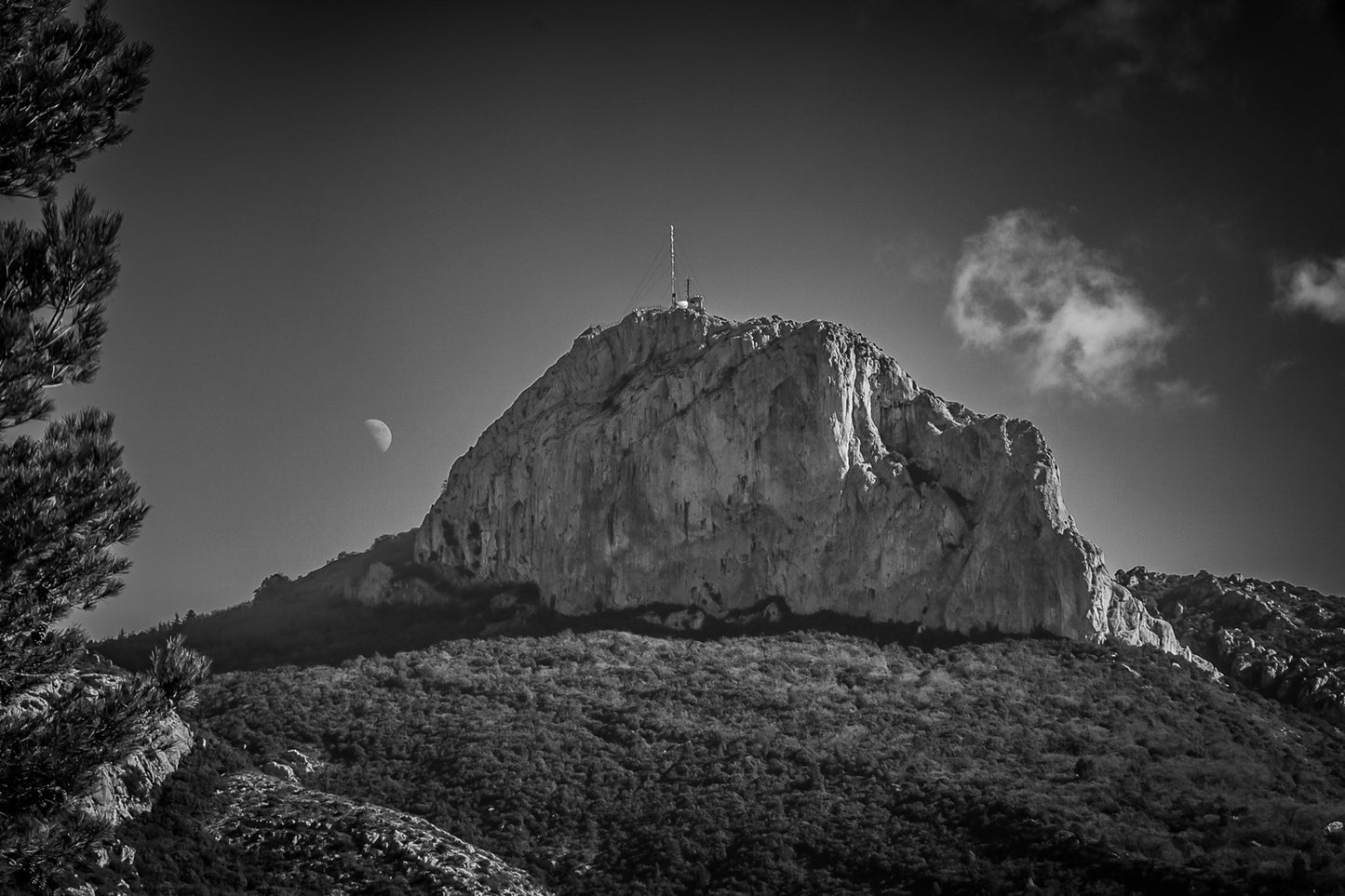 Sommet rocheux de la Sainte-Baume avec antenne au sommet, lune à l’horizon et forêt sombre au premier plan, en noir et blanc.