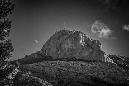 Sommet rocheux de la Sainte-Baume avec antenne au sommet, lune à l’horizon et forêt sombre au premier plan, en noir et blanc.