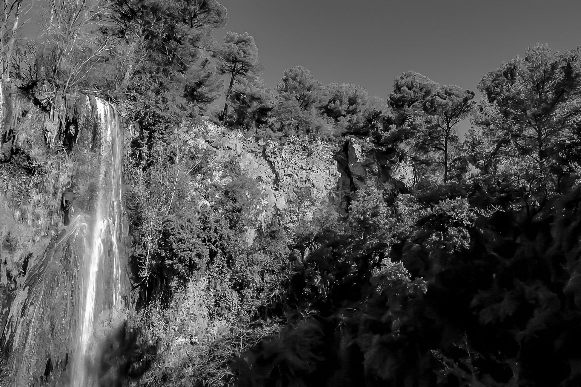 Paroi rocheuse orangée et pins verts dominant la cascade de Sillans sous un ciel bleu intense, version noir et blanc