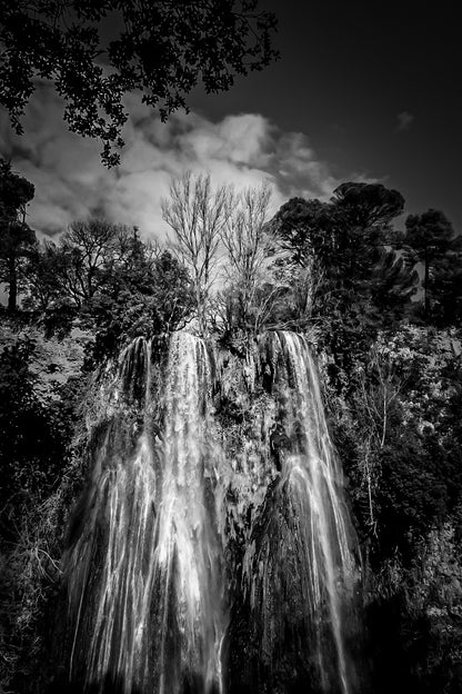 Haute cascade de Sillans se détachant sur un ciel bleu, encadrée par les arbres de la forêt, noir et blanc