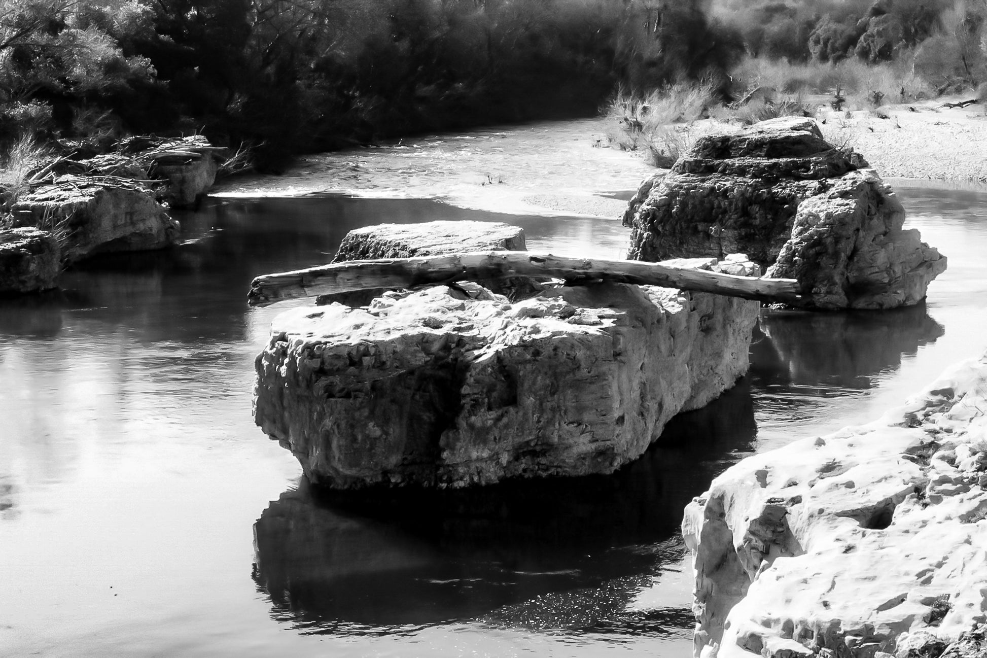 Gros rocher au milieu de la rivière Cèze avec un tronc posé en travers à la cascade du Sautadet, noir et blanc