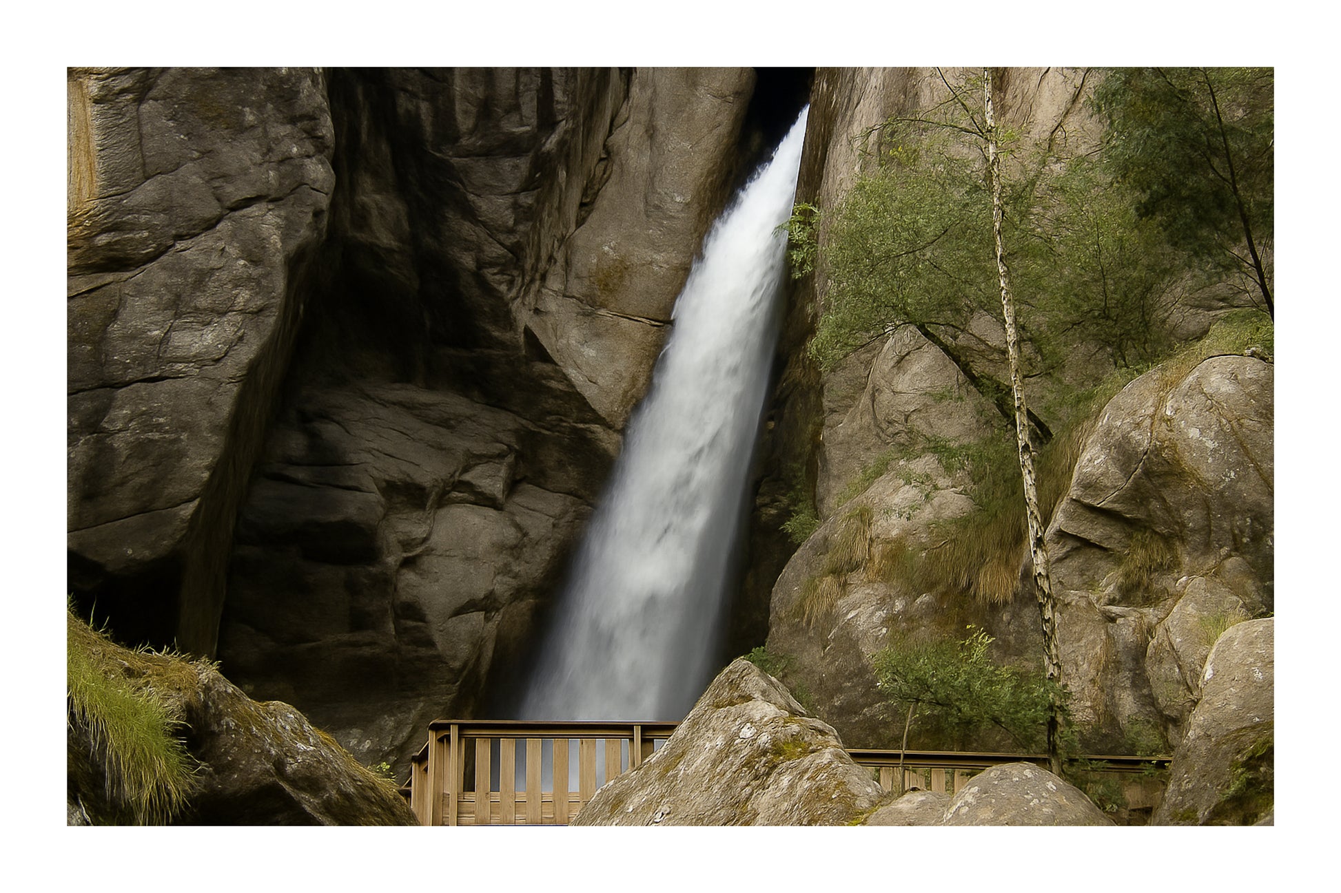 Grande chute d’eau de la cascade du Ray au fond d’une faille rocheuse, passerelle de bois au premier plan, couleur avec bordure