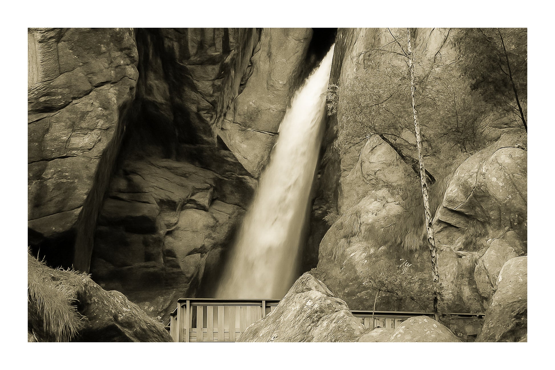 Grande chute d’eau de la cascade du Ray au fond d’une faille rocheuse, passerelle de bois au premier plan, rendu vintage avec bordure