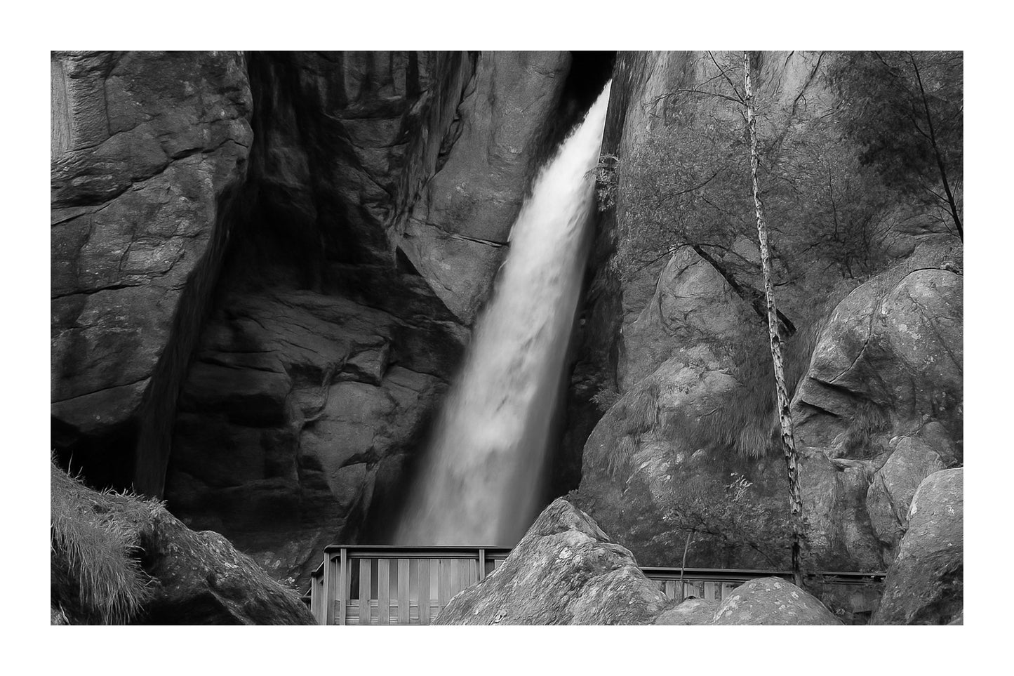 Grande chute d’eau de la cascade du Ray au fond d’une faille rocheuse, passerelle de bois au premier plan, noir et blanc avec bordure