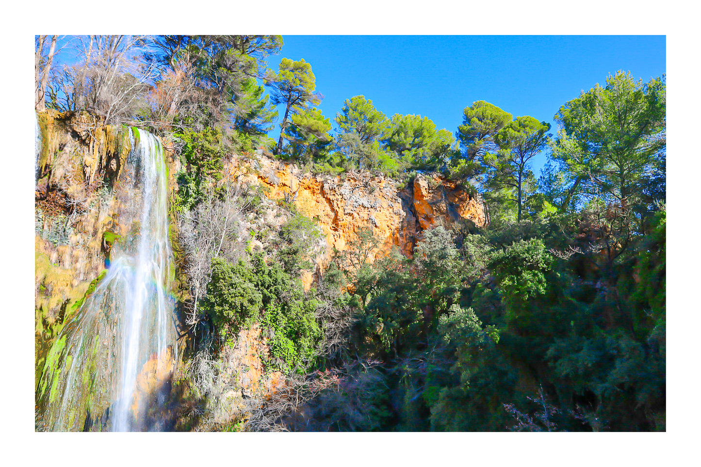 Paroi rocheuse orangée et pins verts dominant la cascade de Sillans sous un ciel bleu intense avec bordure