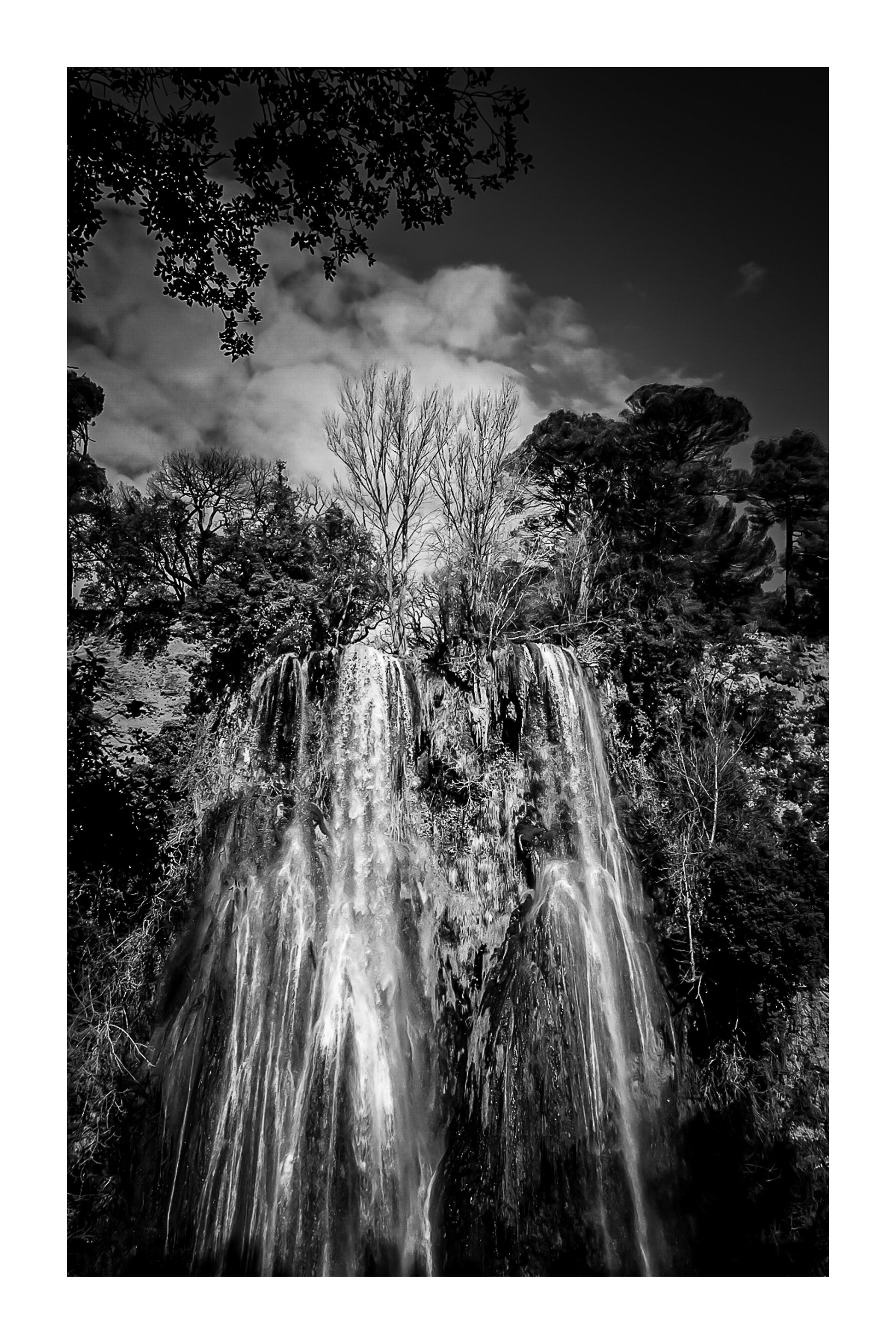 Haute cascade de Sillans se détachant sur un ciel bleu, encadrée par les arbres de la forêt, noir et blanc avec bordure