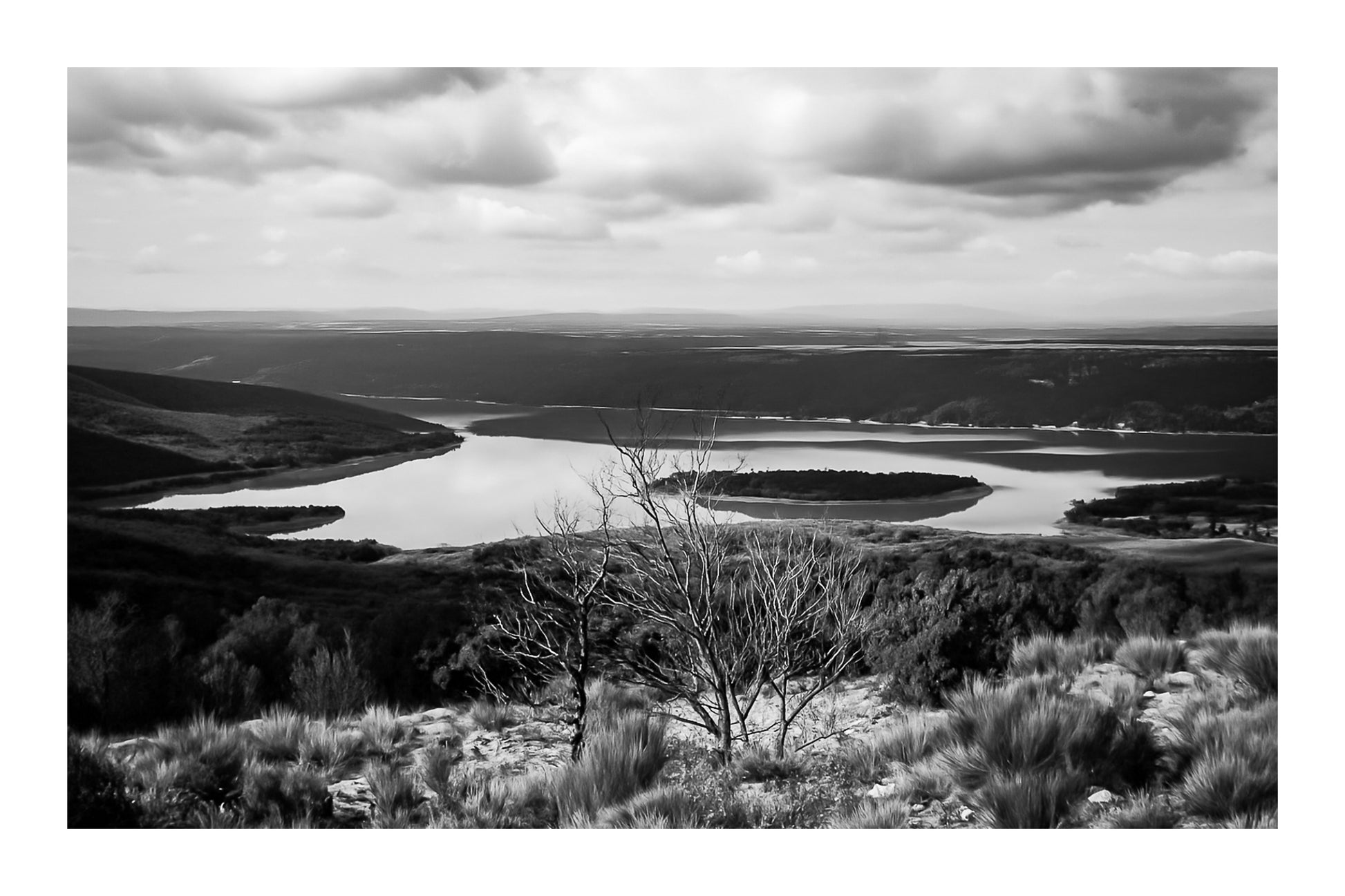 Vue panoramique sur le lac de Sainte-Croix, bras d’eau sinueux et collines provençales sous un ciel nuageux, noir et blanc avec bordure