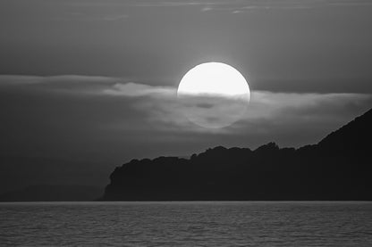 Gros plan sur le soleil rougeoyant partiellement voilé par un nuage au-dessus de la silhouette sombre de Giens, noir et blanc