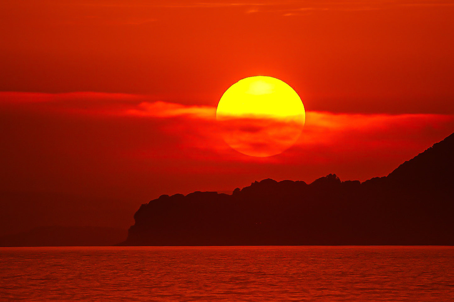 Gros plan sur le soleil rougeoyant partiellement voilé par un nuage au-dessus de la silhouette sombre de Giens