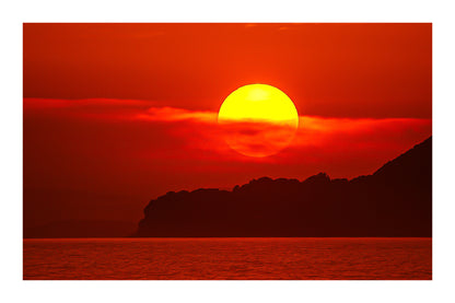 Gros plan sur le soleil rougeoyant partiellement voilé par un nuage au-dessus de la silhouette sombre de Giens avec bordure