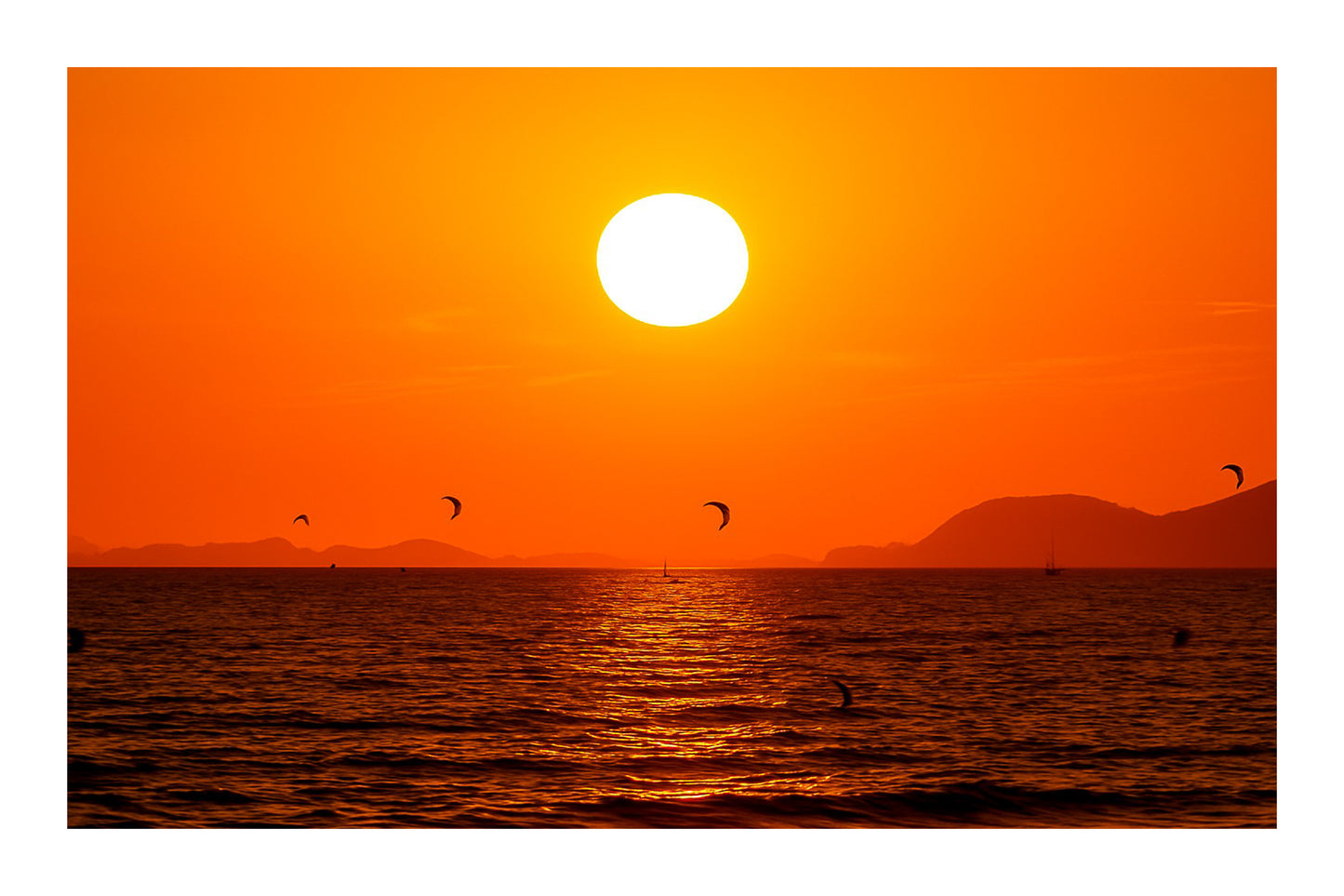 Soleil couchant orange au-dessus de la mer avec des ailes de kitesurf en silhouette à l’Almanarre, avec bordure