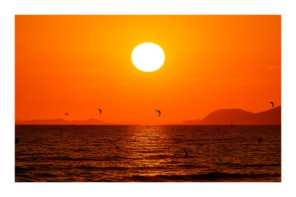 Soleil couchant orange au-dessus de la mer avec des ailes de kitesurf en silhouette à l’Almanarre, avec bordure