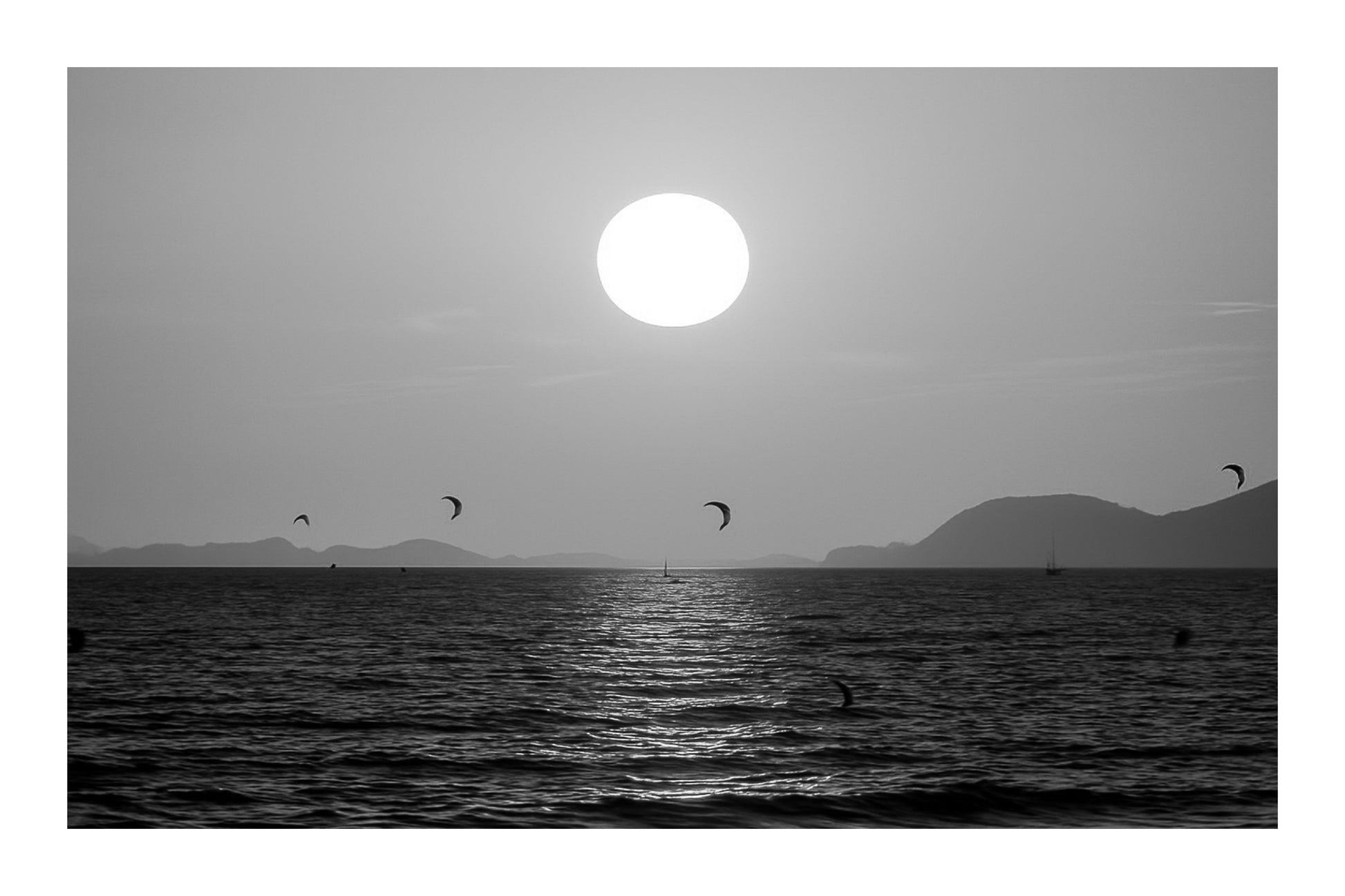Soleil couchant orange au-dessus de la mer avec des ailes de kitesurf en silhouette à l’Almanarre, noir et blanc avec bordure