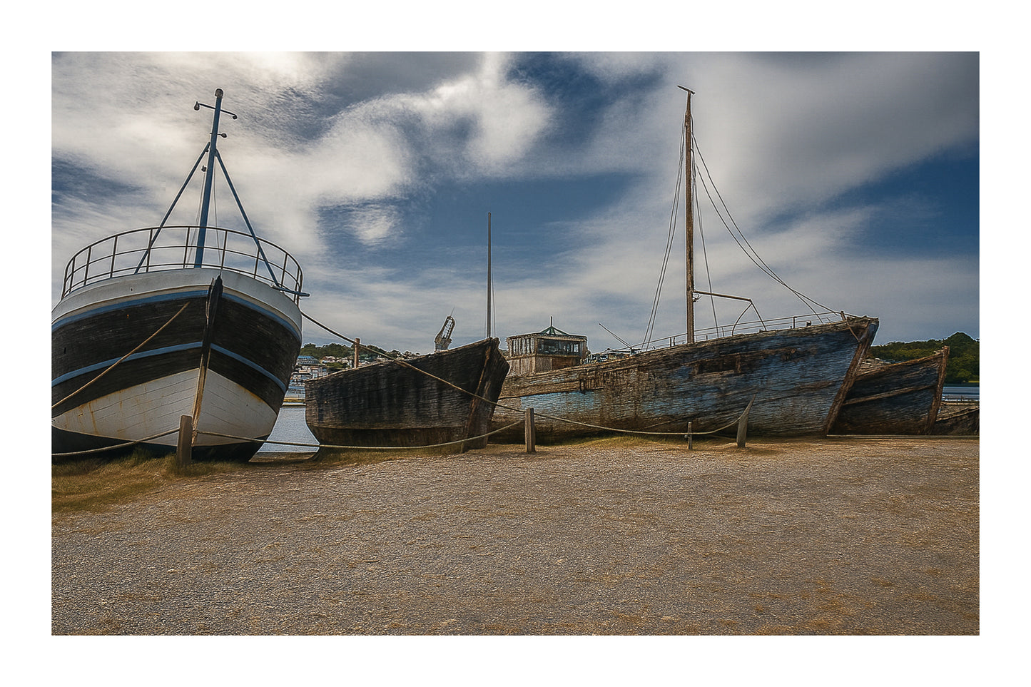 Trois bateaux en bois alignés sur la grève à Camaret, ciel nuageux et port en arrière-plan, avec bordure