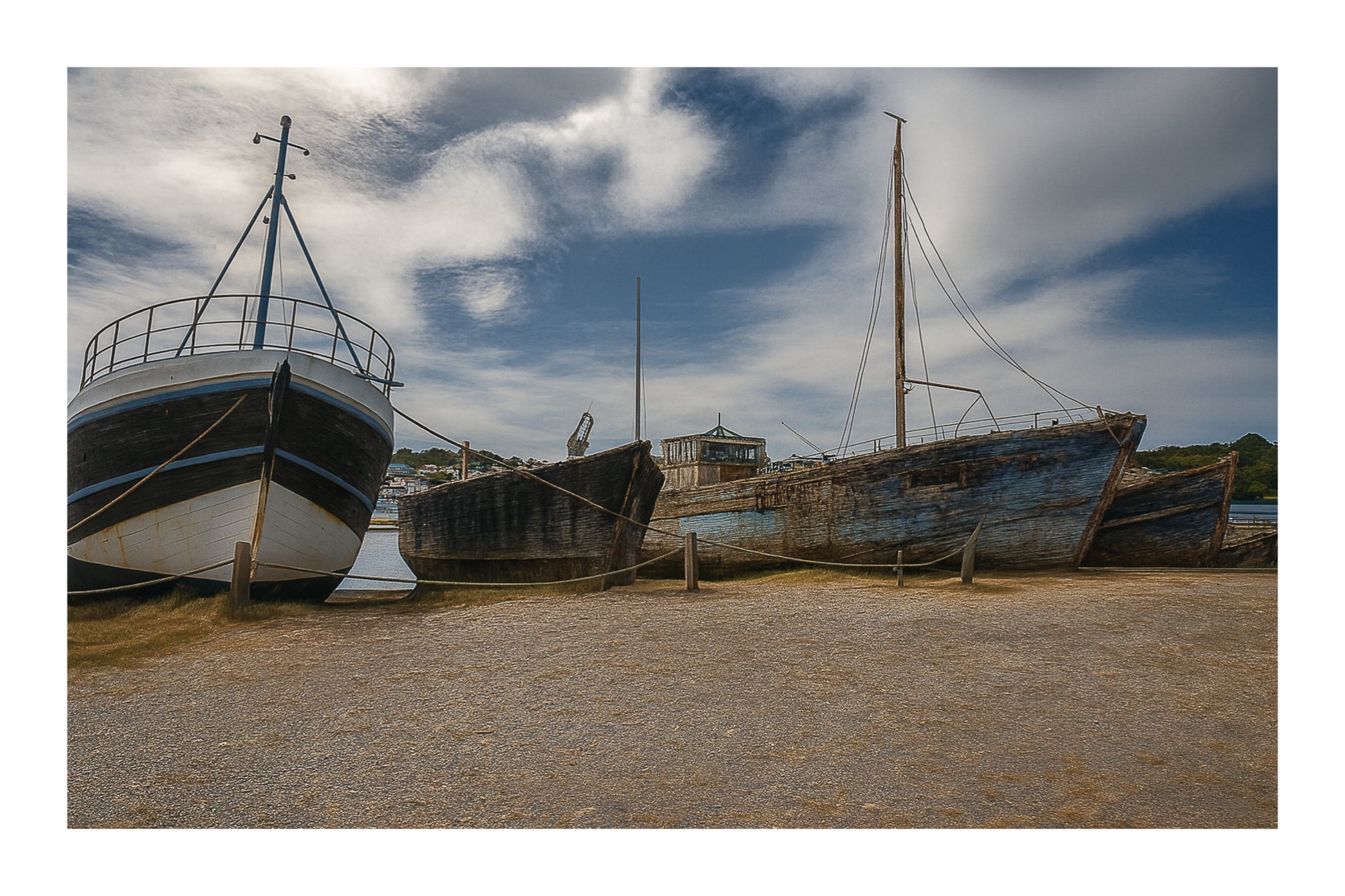 Trois bateaux en bois alignés sur la grève à Camaret, ciel nuageux et port en arrière-plan, avec bordure