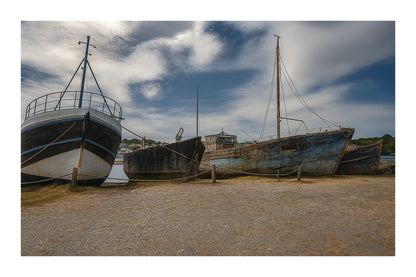 Trois bateaux en bois alignés sur la grève à Camaret, ciel nuageux et port en arrière-plan, avec bordure