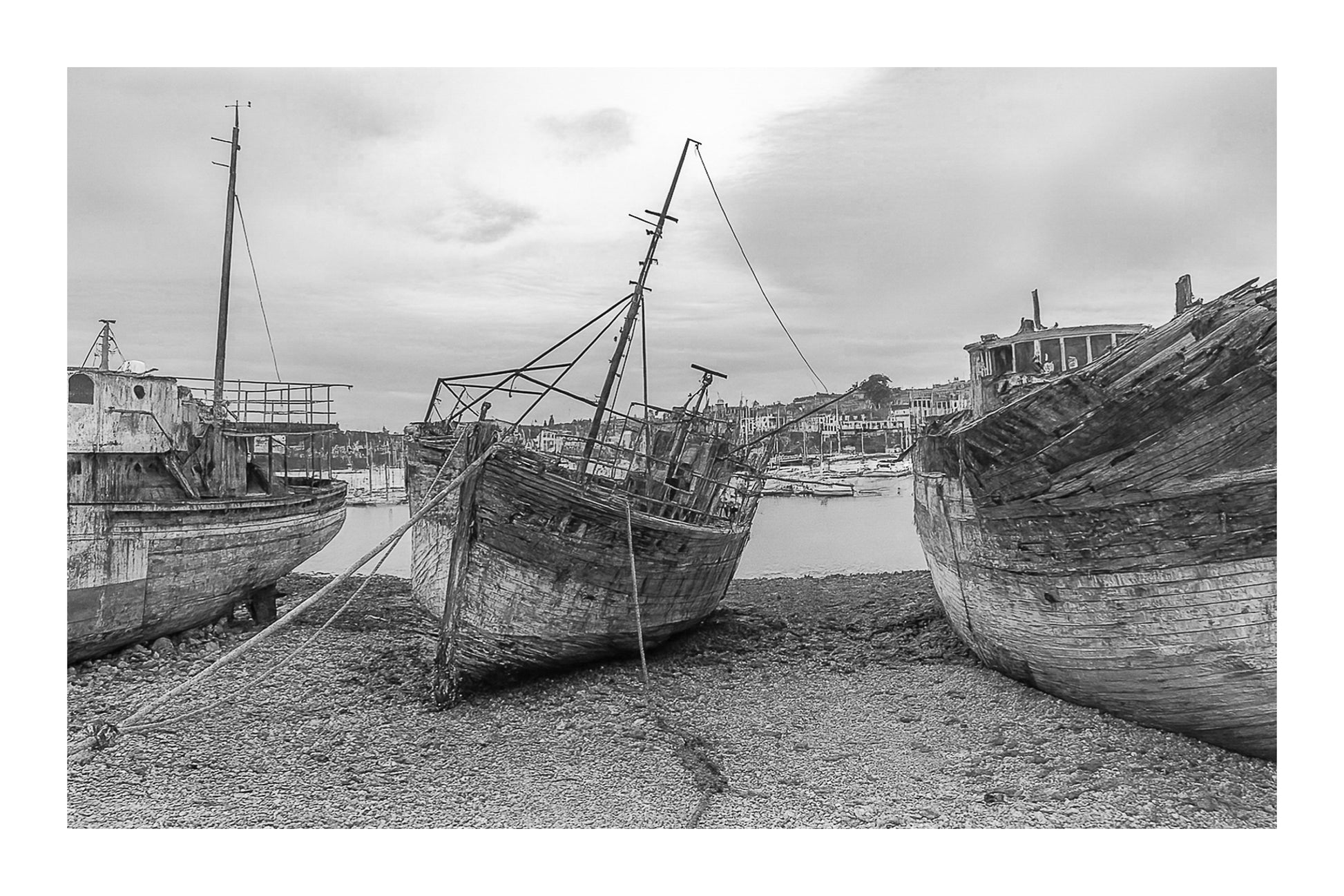 Vieille coque bleue au centre des épaves sur galets, ville de Camaret au fond, noir et blanc avec bordure