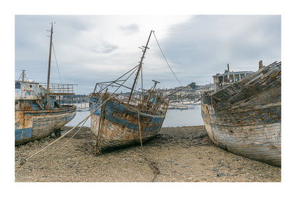 Vieille coque bleue au centre des épaves sur galets, ville de Camaret au fond, avec bordure