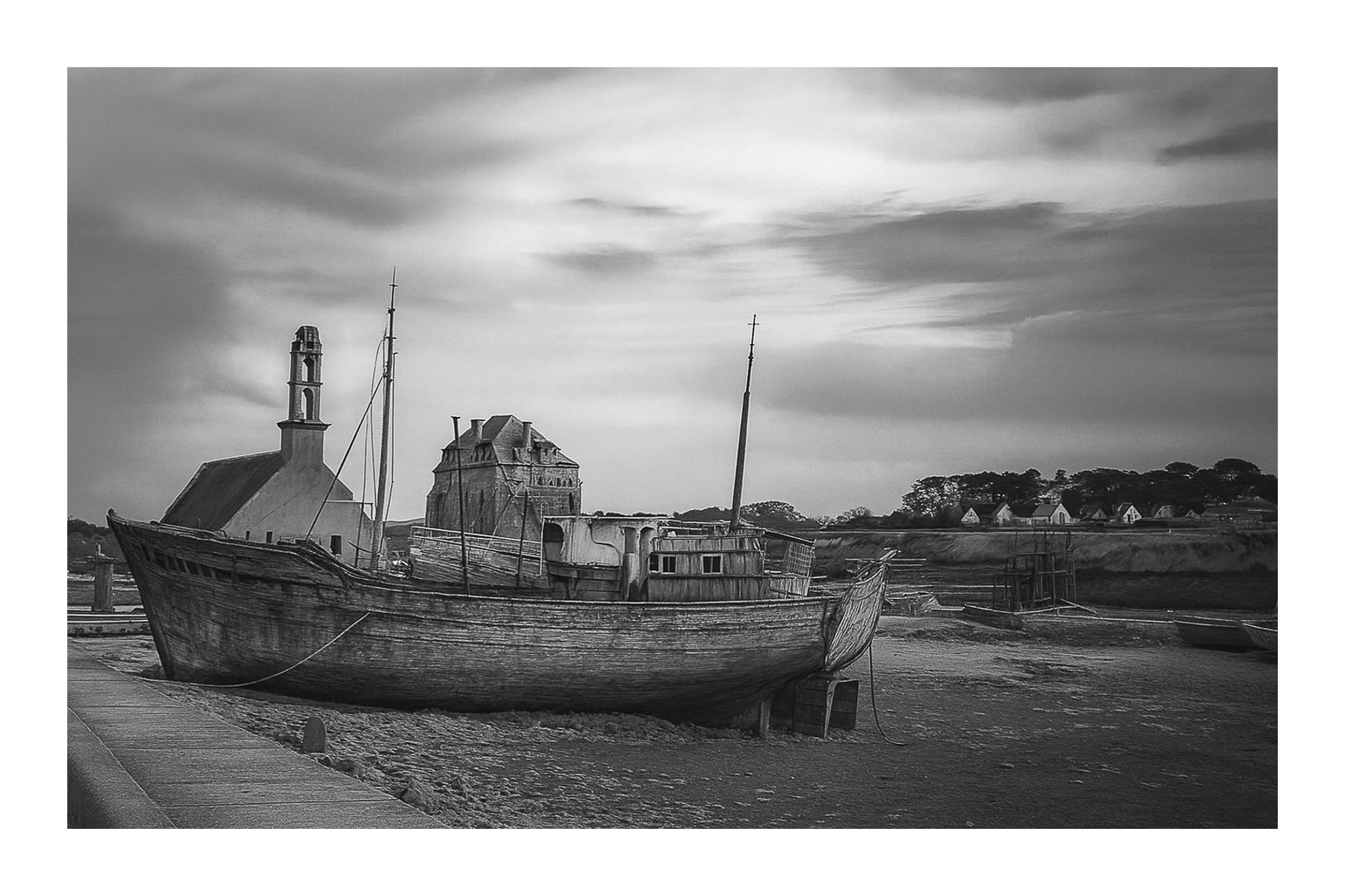 Épave bleue au premier plan devant chapelle et maisons, ciel doré au-dessus du port de Camaret, noir et blanc avec bordure