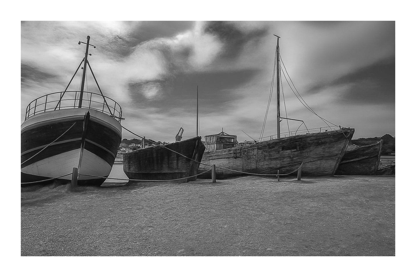 Trois bateaux en bois alignés sur la grève à Camaret, ciel nuageux et port en arrière-plan, noir et blanc avec bordure